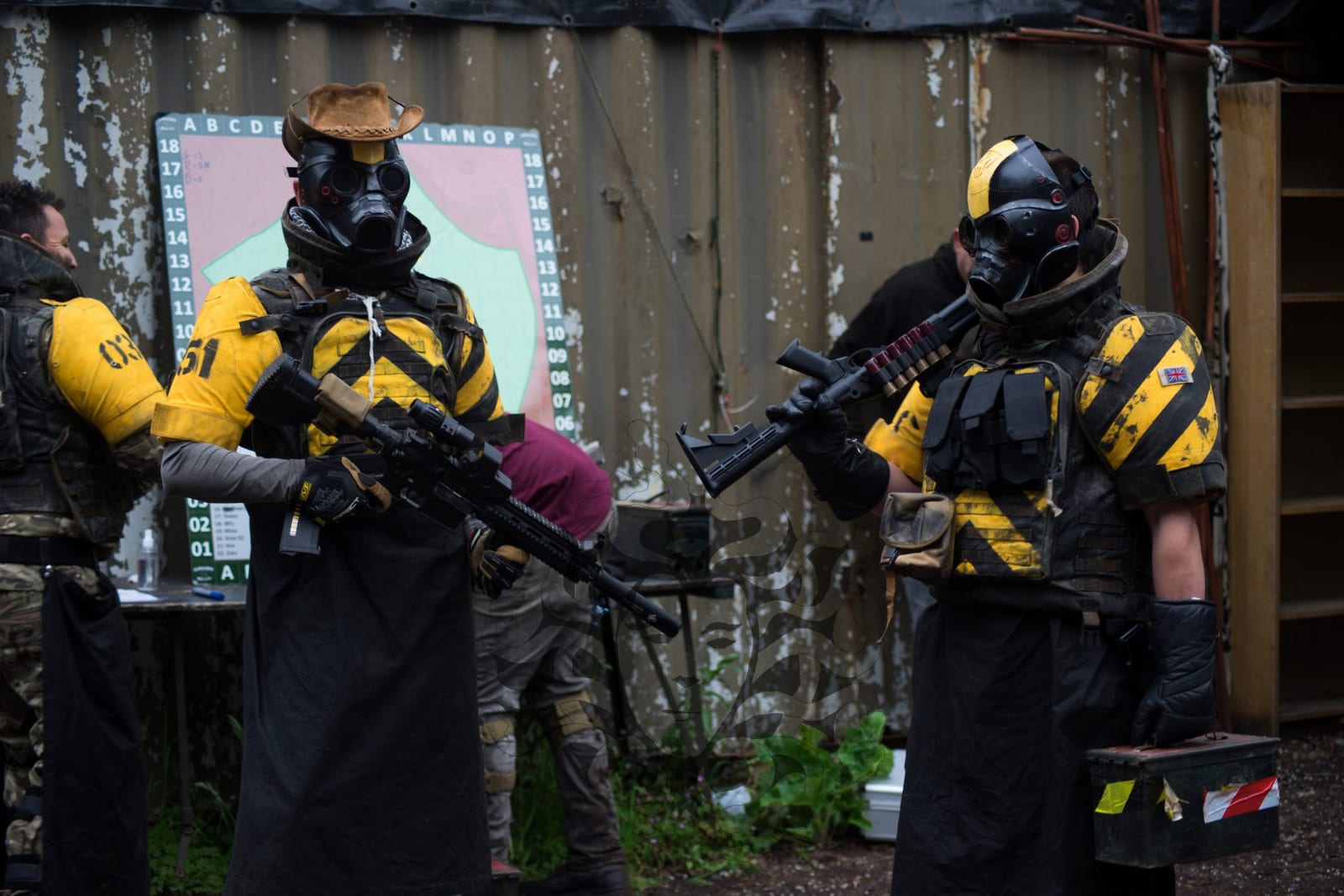 Two airsoft players in yellow and black tactical kit, gas masks and replica rifles at a container-walled UK skirmish site