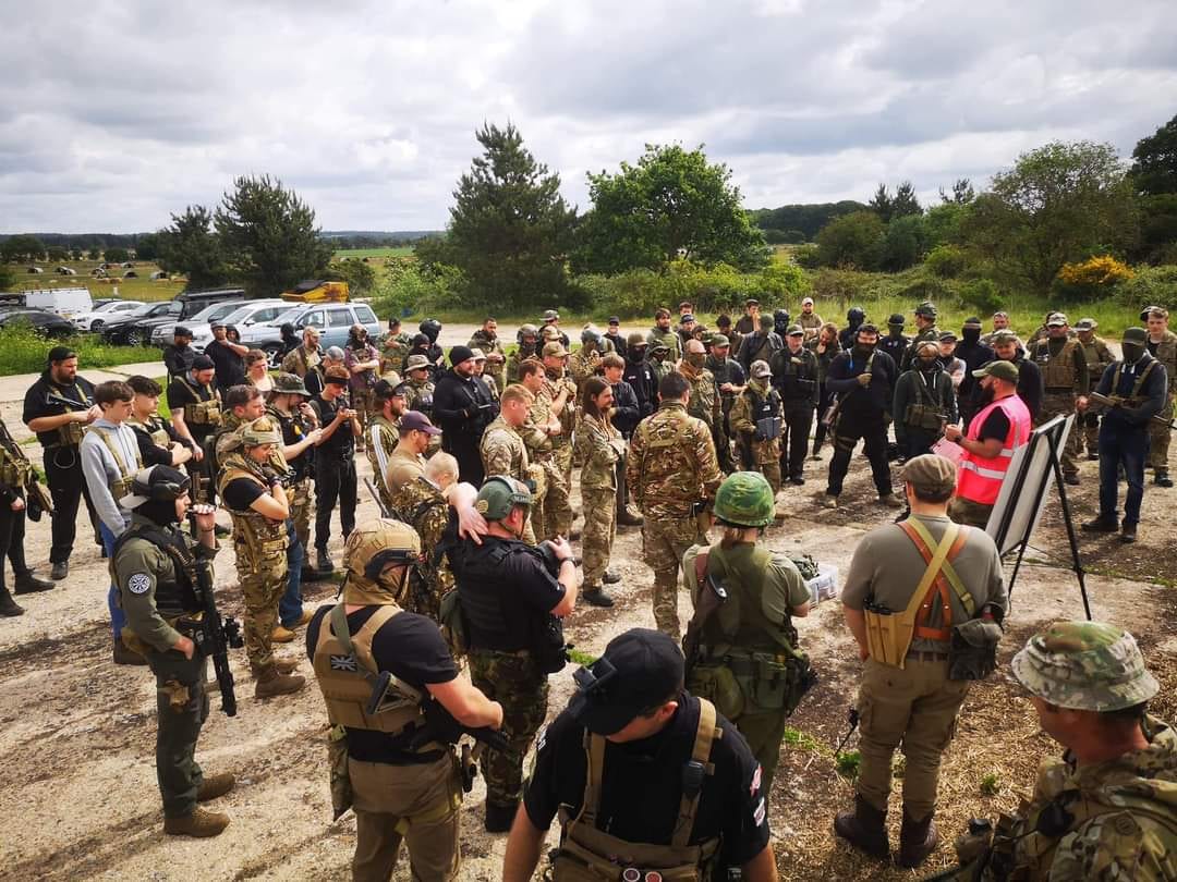 A large crowd of airsoft players in tactical kit gathered for a safe-zone game briefing at a UK skirmish event, with a marshal in a hi-vis vest