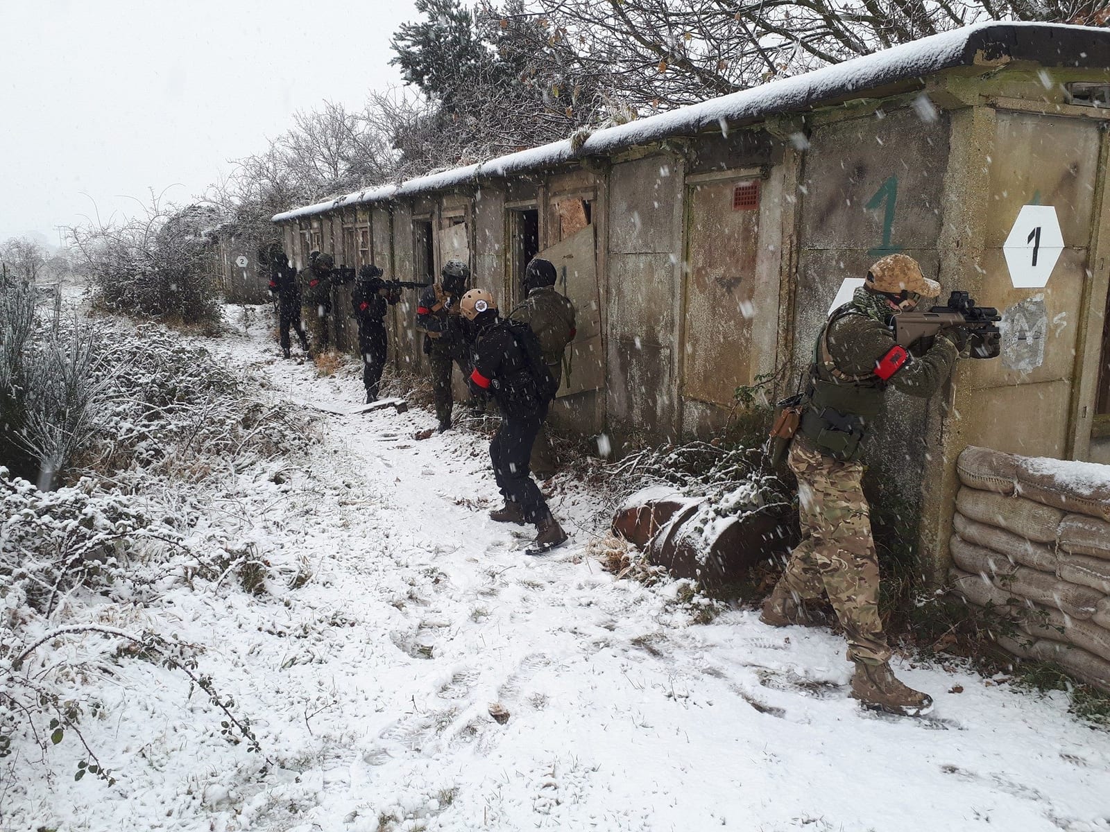 A squad of airsoft players in camouflage advancing along a snow-covered CQB garage block with replica rifles at a UK skirmish site