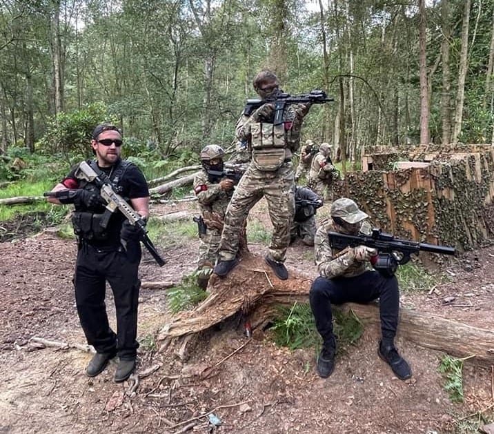 A team of airsoft players in camouflage with replica rifles posed among logs and cover in a UK woodland skirmish site