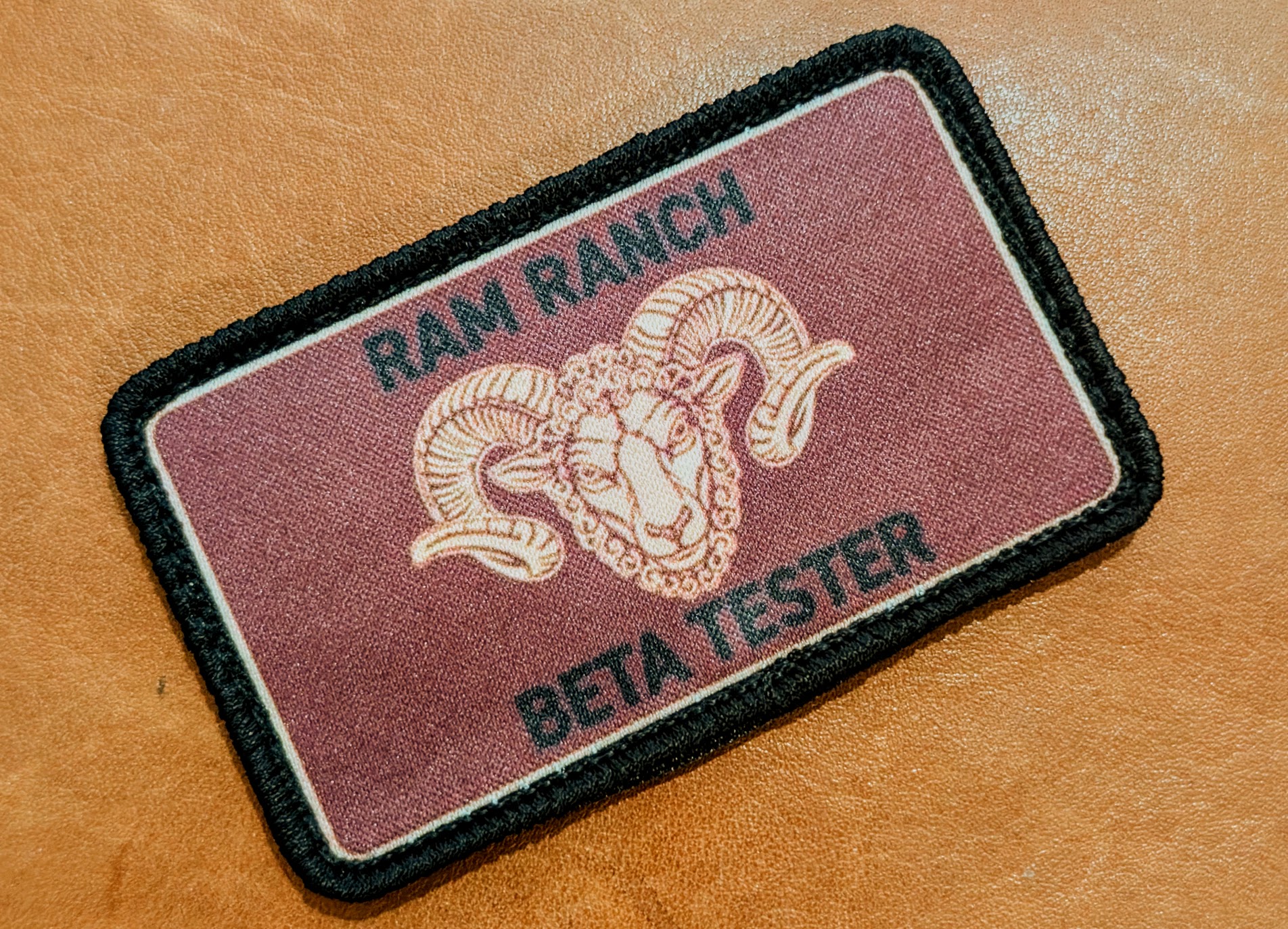The Ram Ranch Beta Tester patch: a maroon embroidered patch reading RAM RANCH BETA TESTER with an orange ram head and a black merrowed border, resting on leather