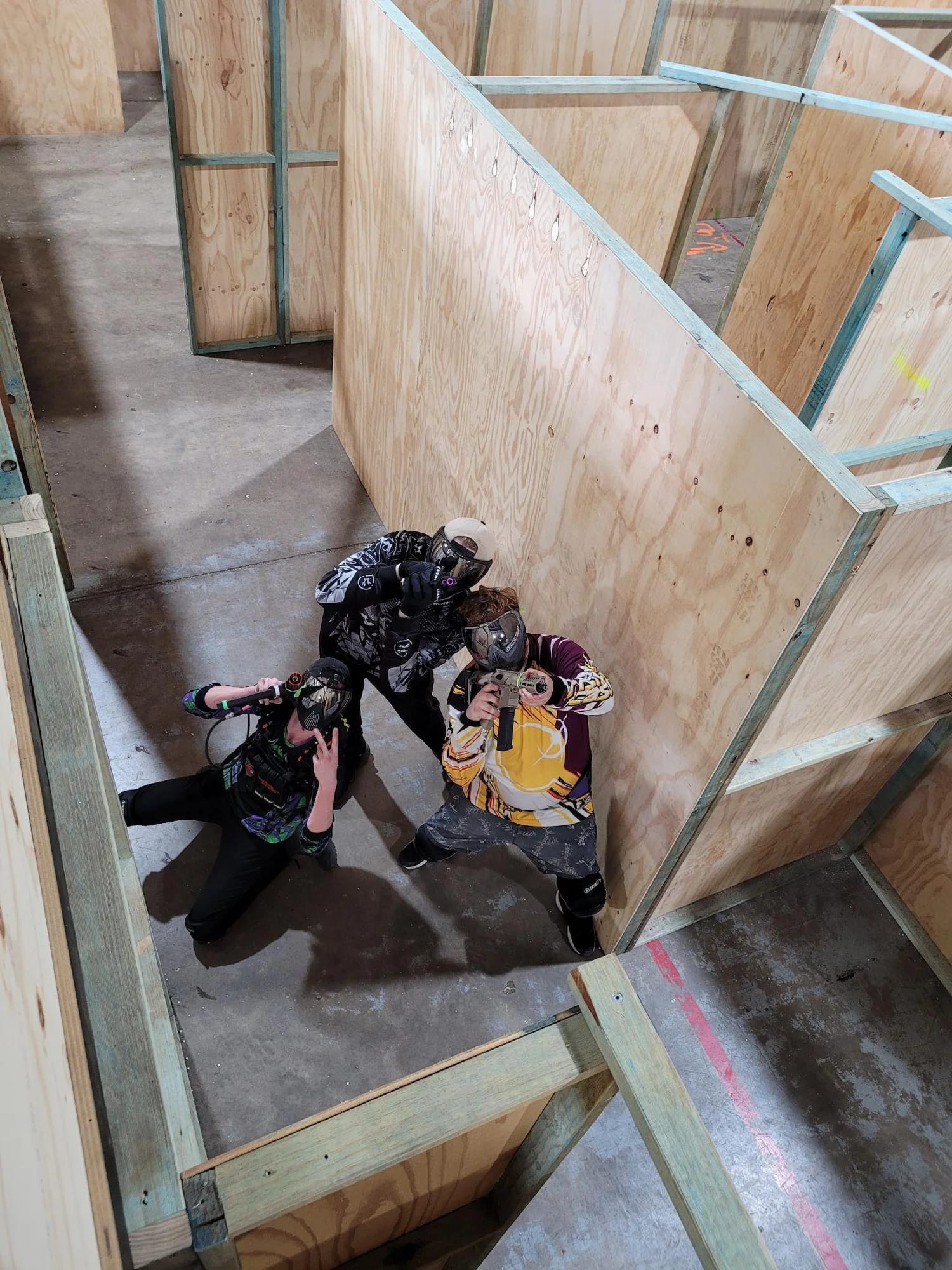 Three gel ball players in bright competition jerseys and face masks holding gel blasters inside the plywood CQB maze at Urban Gel Ball, Queensland